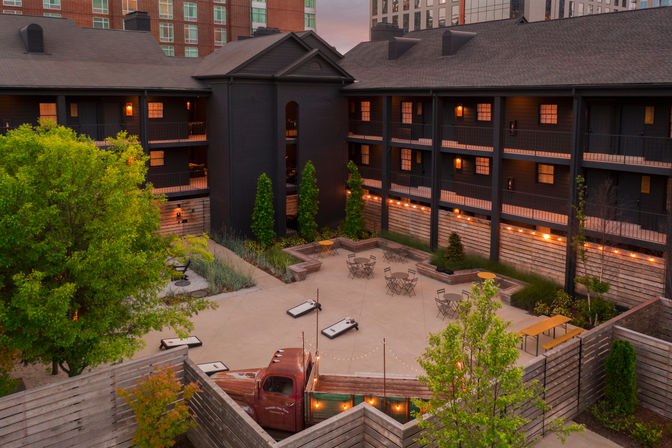 Urban hotel courtyard at dusk with string lights, patio tables, cornhole boards and a vintage truck centerpiece, surrounded by dark wood three-story balconies and city buildings.