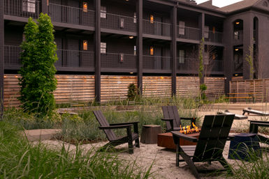 Cozy modern outdoor courtyard with black Adirondack chairs around a rectangular fire pit, sandy gravel, ornamental grasses, wood-panel fence and dark multi-story building with balconies and warm string lights.