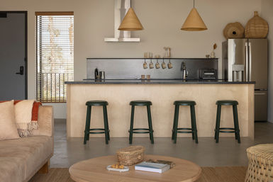 Open-plan neutral-toned living room and kitchen with a light wood island, four dark green bar stools, cone pendant lights, stainless refrigerator, hanging mugs on a dark backsplash, and a coffee table with books and a woven basket.