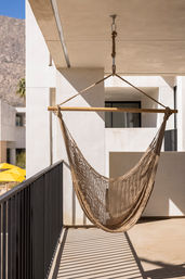 Woven macramé hammock chair hanging on a modern sunlit balcony with black metal railing, mountain backdrop and yellow pool umbrellas below