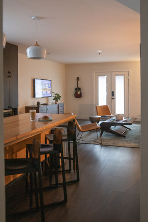 Cozy modern open-plan apartment interior with a wooden kitchen island and black bar stools, pendant light, latte and pastry on the counter, wall-mounted TV, acoustic guitar on the wall, leather lounge chairs and a low coffee table by glass French doors.