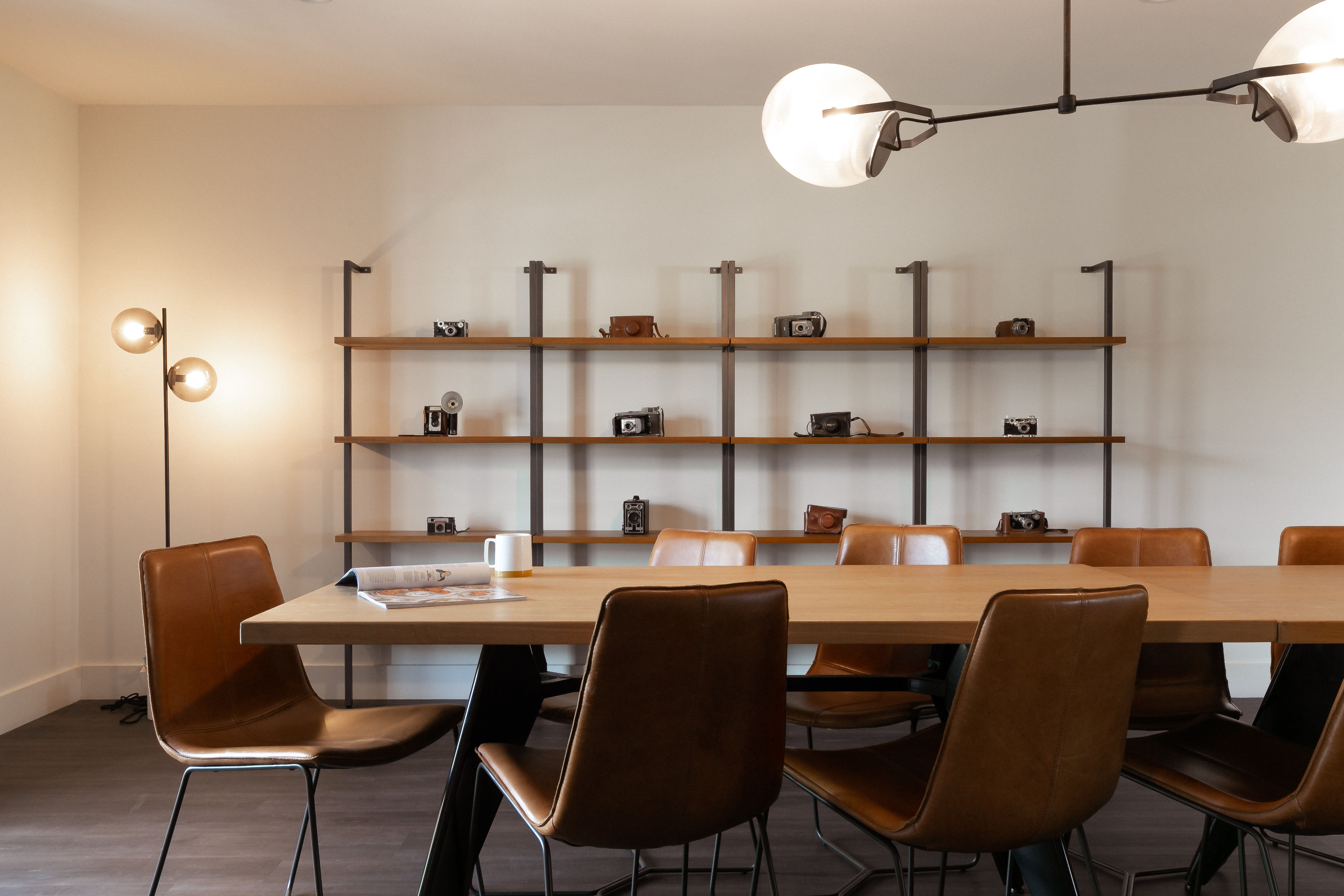 Modern meeting room with a long wooden table and brown leather chairs, wall shelves showcasing vintage cameras, and warm globe pendant and floor lamps.
