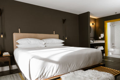 Sleek modern hotel bedroom with king-size bed in crisp white linens, tan leather headboard against a dark gray accent wall, hanging pendant lights, wooden bench with fluffy white rug, and bright yellow-trimmed en-suite bathroom visible