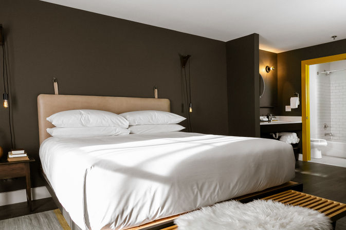 Sleek modern hotel bedroom with king-size bed in crisp white linens, tan leather headboard against a dark gray accent wall, hanging pendant lights, wooden bench with fluffy white rug, and bright yellow-trimmed en-suite bathroom visible
