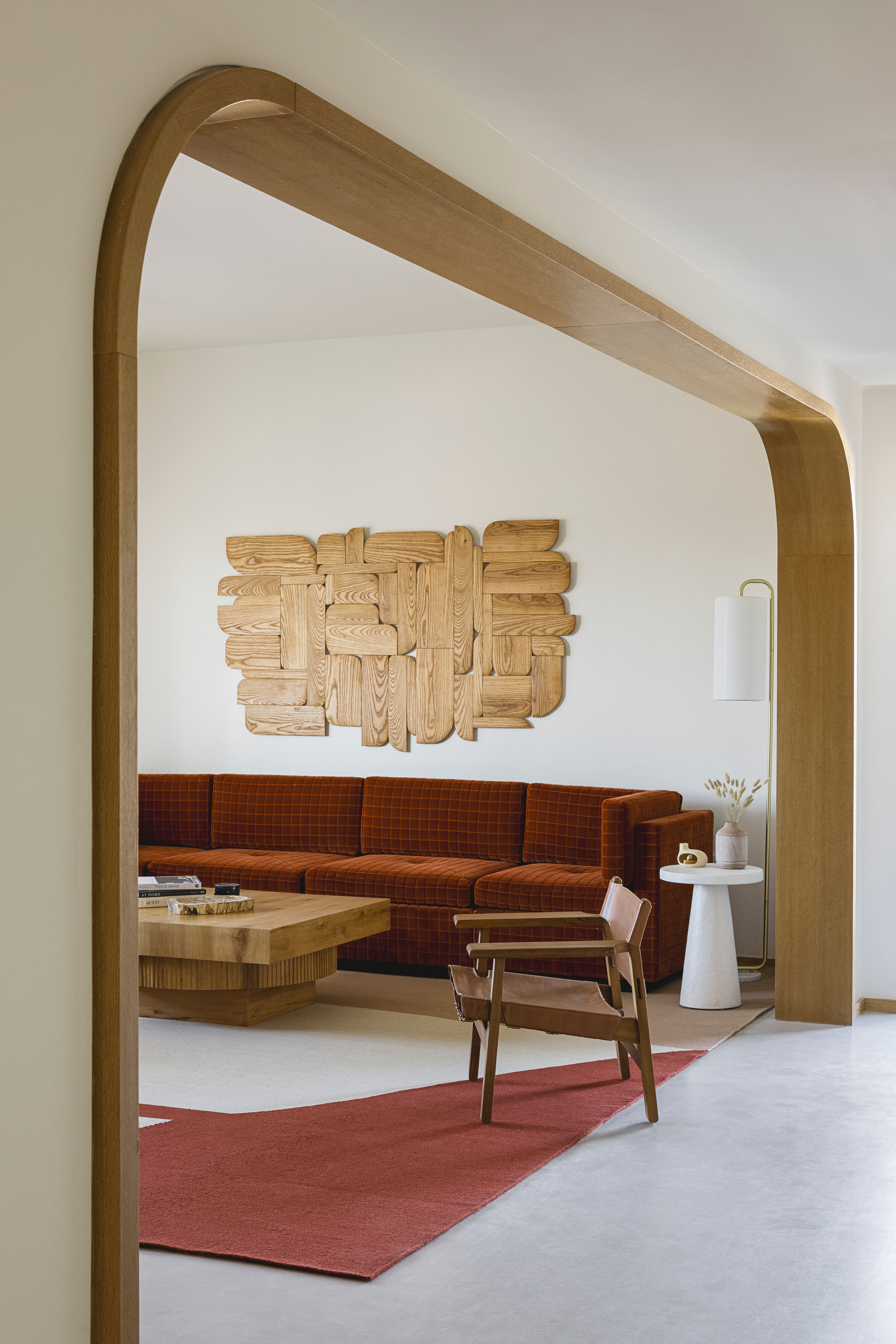 Modern living room seen through an arched oak doorway featuring a rust-colored velvet sectional, geometric wooden wall art, chunky oak coffee table, leather sling chair and red area rug on neutral floors.