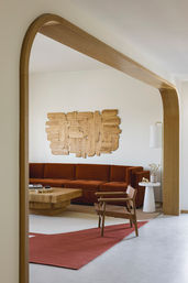 Modern living room seen through an arched oak doorway featuring a rust-colored velvet sectional, geometric wooden wall art, chunky oak coffee table, leather sling chair and red area rug on neutral floors.
