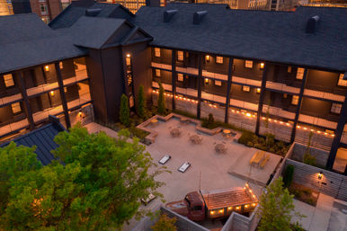 Dusk aerial view of a cozy downtown urban courtyard surrounded by multi-story buildings with lit balconies, string lights, patio tables, cornhole boards and a vintage truck converted into a lit seating feature.