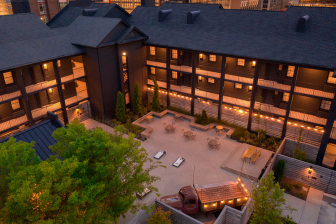 Dusk aerial view of a cozy downtown urban courtyard surrounded by multi-story buildings with lit balconies, string lights, patio tables, cornhole boards and a vintage truck converted into a lit seating feature.
