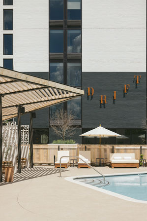 Sunny urban hotel pool deck with wooden slatted pergola casting striped shadows, cushioned lounge chairs and umbrella beside a curved swimming pool and modern black-and-white building facade.