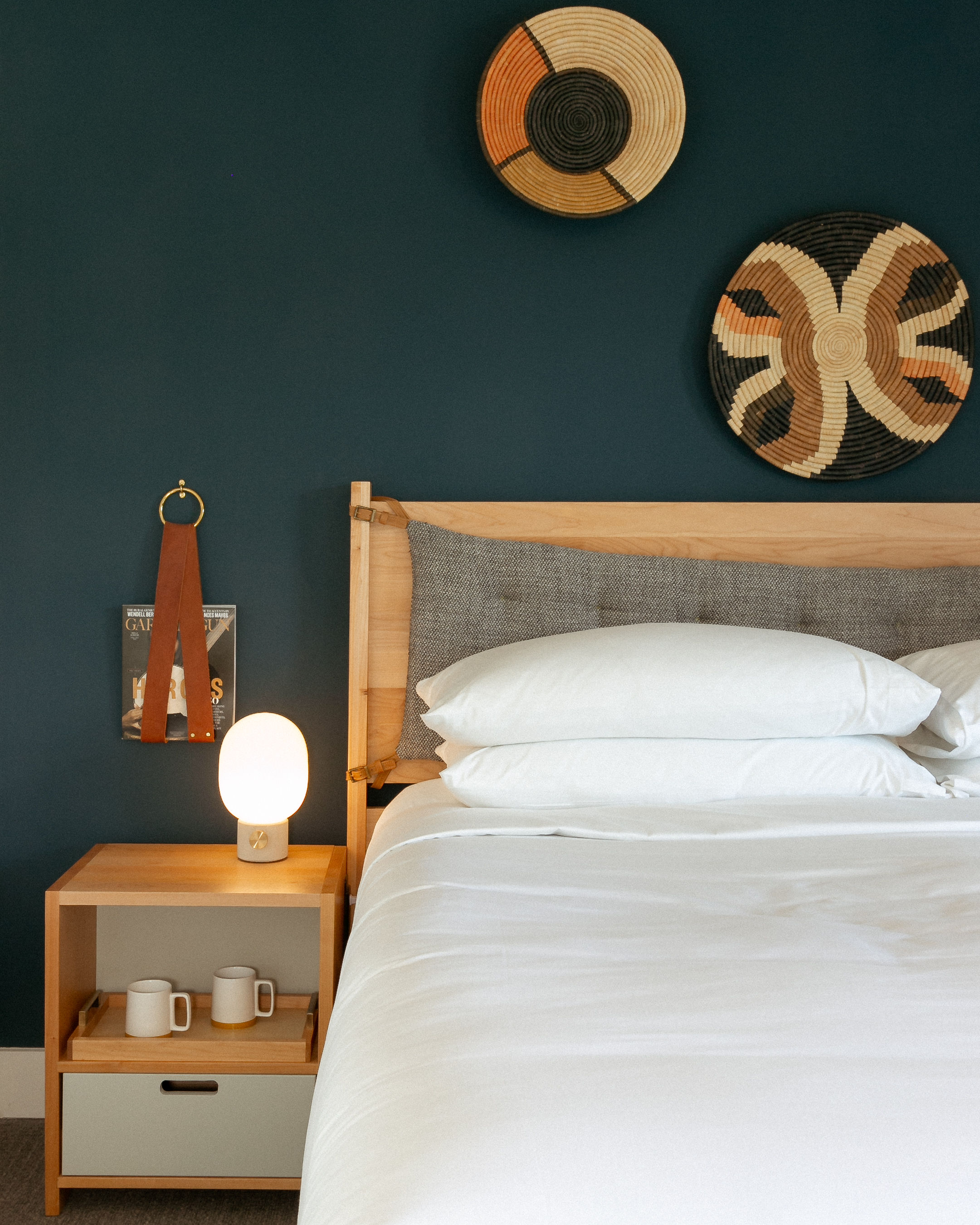 Cozy modern bedroom with a navy accent wall, light wood bed and headboard, crisp white bedding, woven decorative wall baskets, and a wooden nightstand holding a glowing orb lamp and two ceramic mugs.