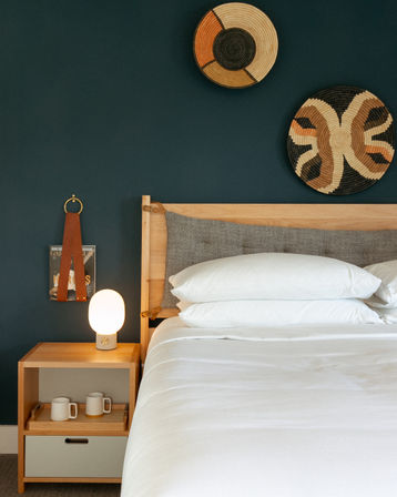 Cozy modern bedroom with a navy accent wall, light wood bed and headboard, crisp white bedding, woven decorative wall baskets, and a wooden nightstand holding a glowing orb lamp and two ceramic mugs.