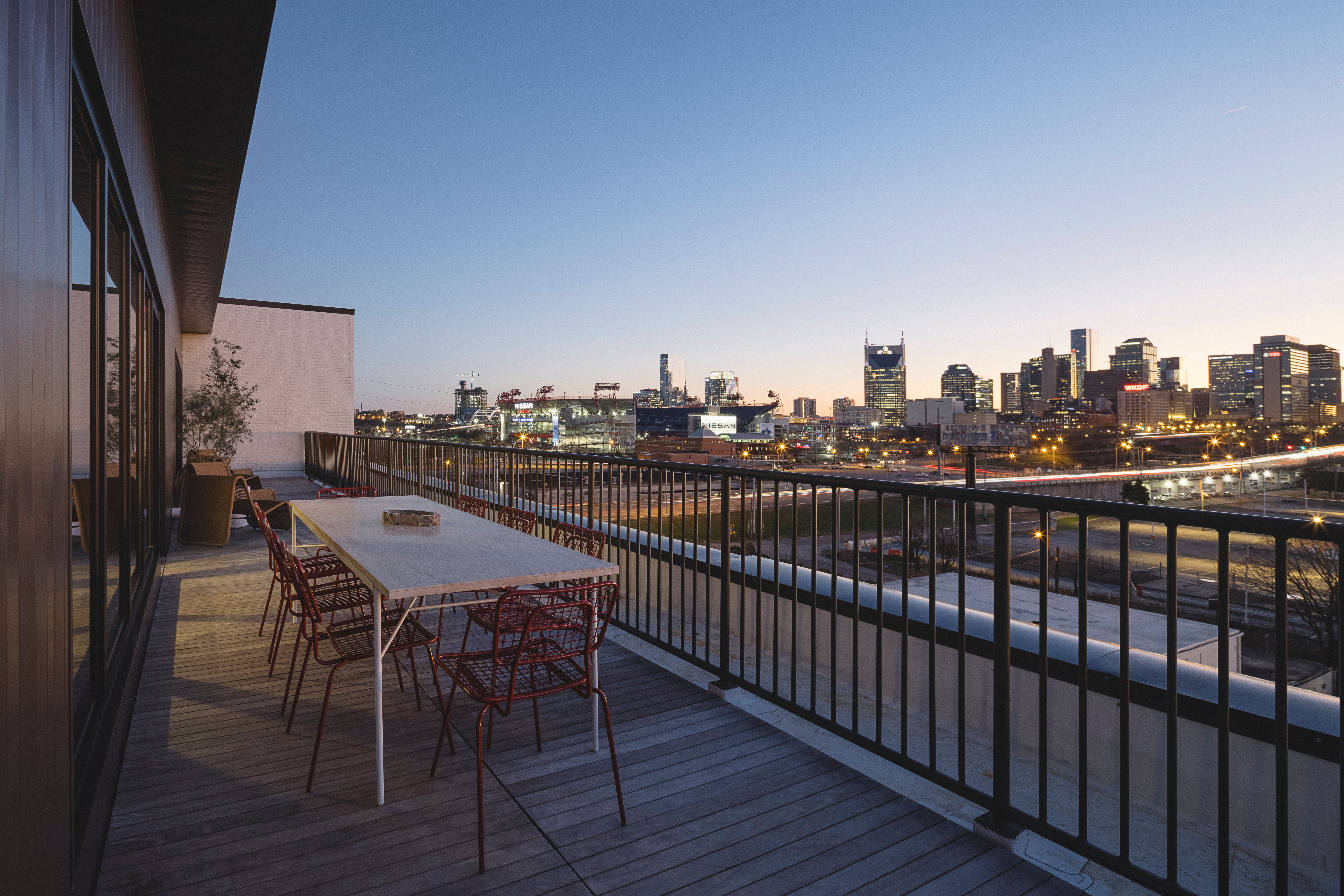 Rooftop terrace with long dining table and red wire chairs on a wooden deck, metal railing framing an illuminated urban skyline and highways at dusk.