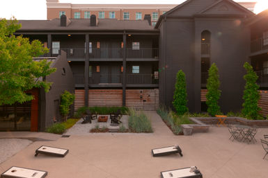 City courtyard at sunset framed by a dark-painted three-story building with balconies, cornhole boards, a fire pit, patio tables and planted greenery.