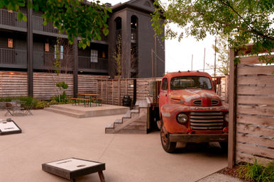 Outdoor urban courtyard patio with a rusty vintage red pickup truck by a wooden fence, picnic table on a raised concrete platform, cornhole boards, string lights and a dark multi-story building in the background.