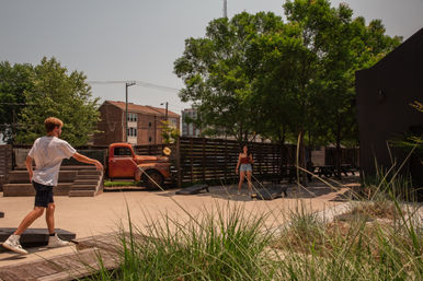 Outdoor urban courtyard with two people playing cornhole under large shade trees, picnic tables, ornamental grasses, and a rusted vintage pickup truck parked by a wooden fence.