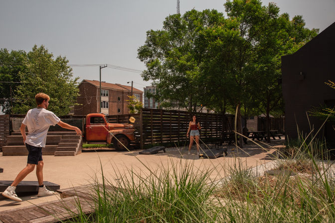 Outdoor urban courtyard with two people playing cornhole under large shade trees, picnic tables, ornamental grasses, and a rusted vintage pickup truck parked by a wooden fence.