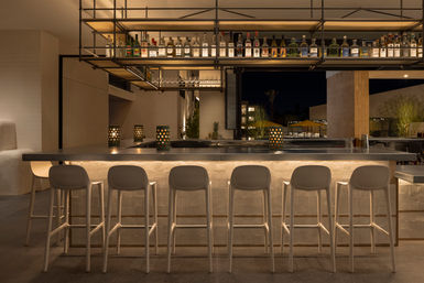 Sleek outdoor hotel bar at night with an illuminated stone counter, hanging shelves of liquor bottles, six white bar stools and warm patio lighting