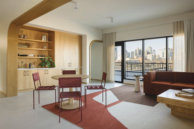 Sunlit modern open-plan apartment living room with round dining table and red woven chairs, built-in light wood shelving, rust-and-cream rug, sofa and balcony overlooking downtown skyline