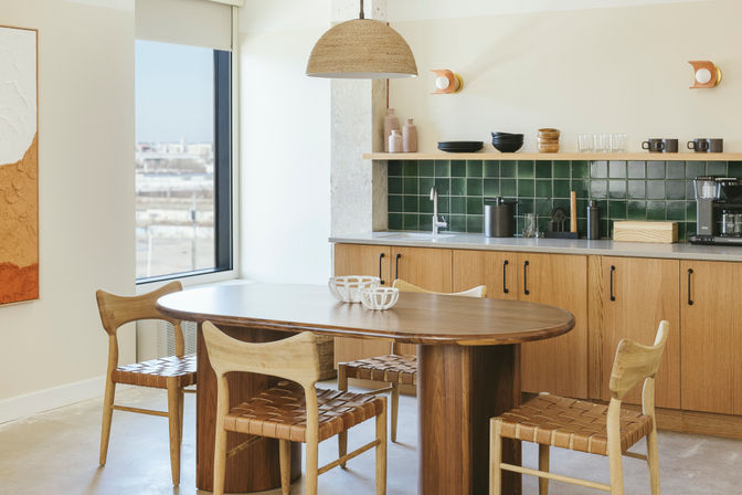 Sunlit Scandi-modern kitchen dining area with oval walnut table, woven leather chairs, green tile backsplash, wood cabinets, rattan pendant and large window view.