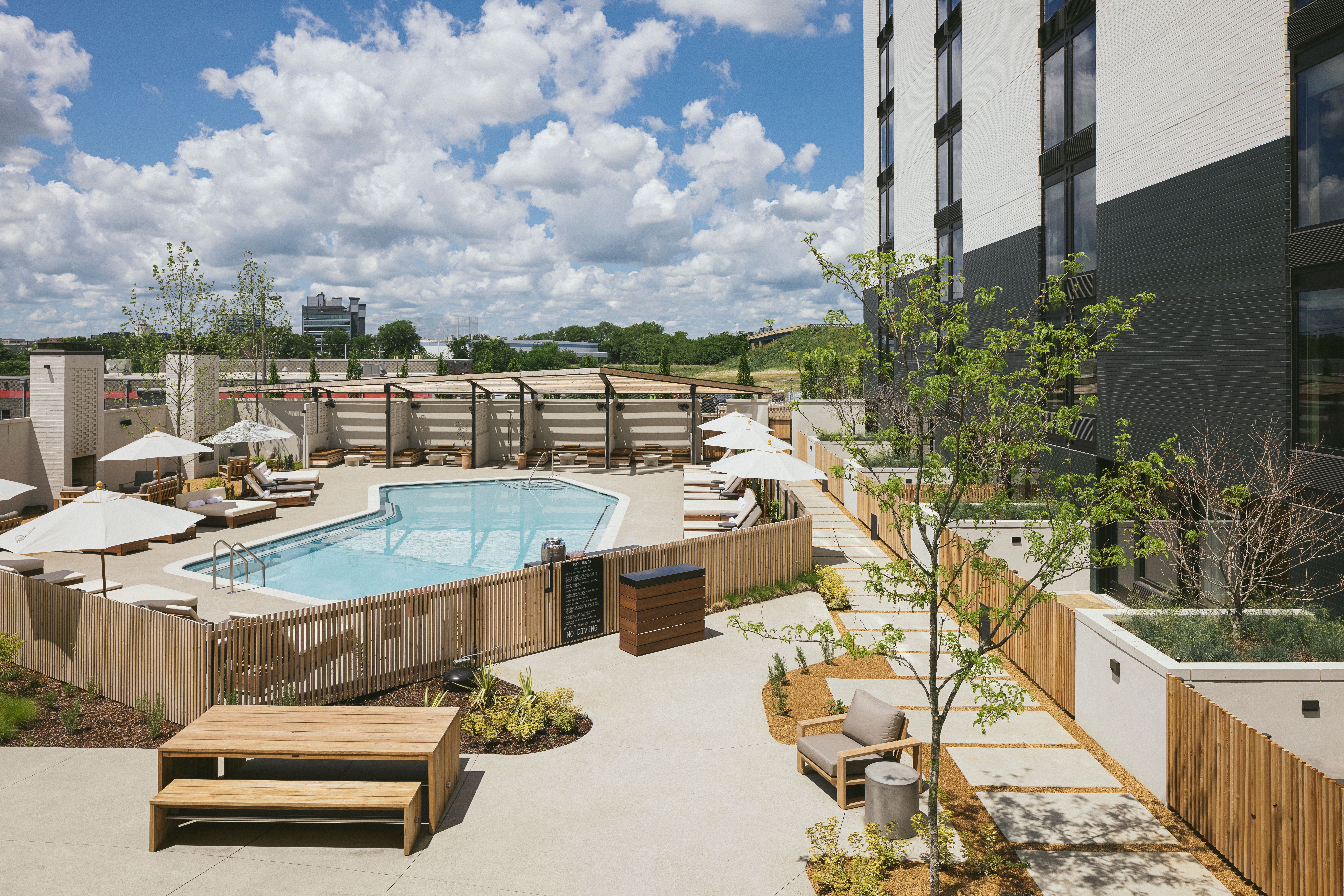 Rooftop pool terrace in an urban setting with a clear blue pool, teak loungers, white umbrellas and cabanas beside a modern high-rise, wooden fencing and city skyline under puffy clouds.