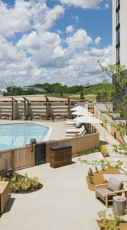 Rooftop pool terrace in an urban setting with a clear blue pool, teak loungers, white umbrellas and cabanas beside a modern high-rise, wooden fencing and city skyline under puffy clouds.