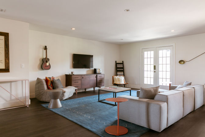 Bright modern living room interior with light gray sectional sofa, blue area rug, hardwood floors, wooden media console with wall-mounted TV, acoustic guitar on the wall, French doors and minimalist decor.