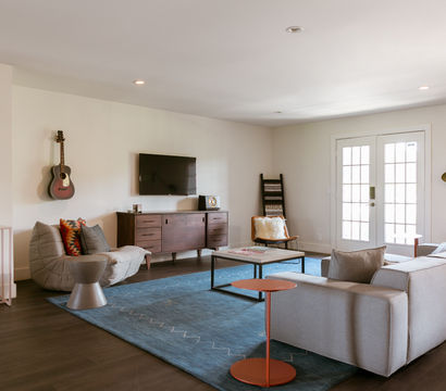 Bright modern living room interior with light gray sectional sofa, blue area rug, hardwood floors, wooden media console with wall-mounted TV, acoustic guitar on the wall, French doors and minimalist decor.