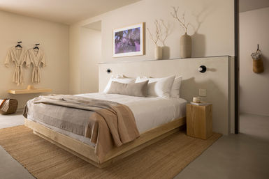 Minimalist modern bedroom with neutral palette, wooden platform bed dressed in layered beige linens and pillows, woven jute rug, wall-mounted robes, and ceramic vases for a calm, natural look.