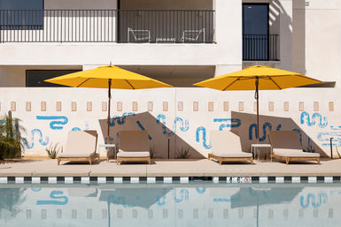 Bright poolside scene: four beige lounge chairs under two yellow umbrellas, blue abstract mural on a modern stucco wall and calm reflecting water.