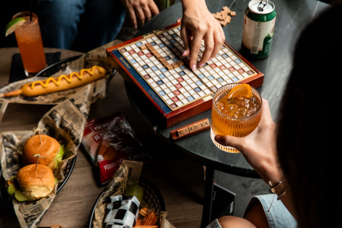 Casual game night at a bar table with hands playing Scrabble surrounded by an orange cocktail, beer can, hot dog, slider burgers and chips
