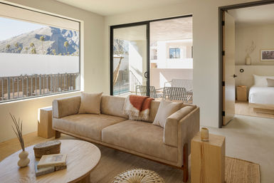 Sunlit modern suite living room with tan suede sofa and throw, round wood coffee table, sliding glass door to balcony with chairs, desert mountain and palm-tree view