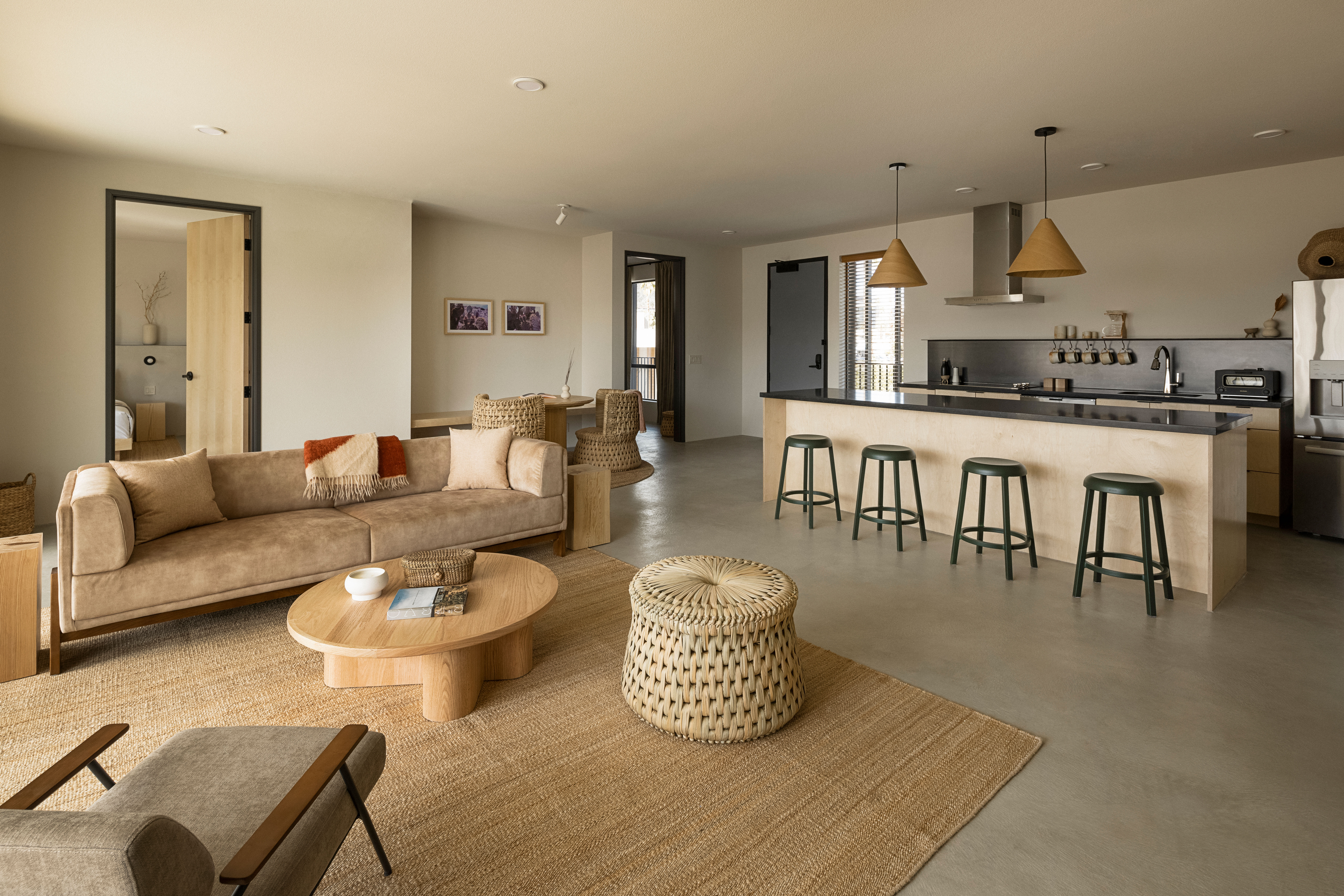 Cozy neutral-toned open-plan living room and kitchen with a beige suede sofa, low round wooden coffee table, woven ottoman on a jute rug, and a light-wood kitchen island with black countertop and four green bar stools under cone pendant lights.