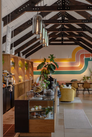 Cozy modern cafe interior with exposed dark wooden beams, hanging glass pendant lights, retro curved wall mural, pastry display and potted plants near lounge seating.