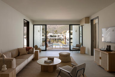 Sunlit modern neutral living room in a resort suite — beige sofa, wooden coffee table and woven pouf, sliding glass doors opening to a balcony overlooking a pool, palm trees and desert mountains