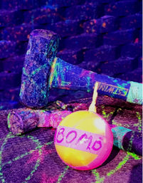 Glow-painted sledgehammers and a neon pink-yellow ball labeled 'BOMB' resting on a paint-splattered floor under blacklight/UV lighting.
