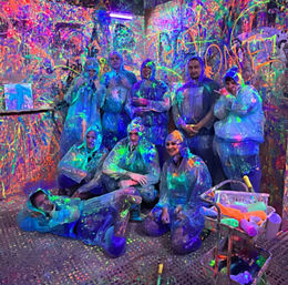 Group of people in disposable coveralls posing in a UV-lit neon paint splatter room, walls and floor covered in colorful fluorescent paint with rollers and a paint cart in the foreground