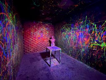 Blacklight neon splatter art room with a paint-covered table centered and a torso bust against the back wall, walls and floor dripped with fluorescent colors.