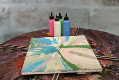 Vibrant acrylic pour canvas on a paint-splattered rotating table with pink, white, green and blue squeeze bottles and a blurred studio wall background.