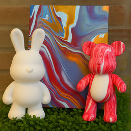 White rabbit vinyl toy and red marbled bear art toy flanking a vibrant abstract marbled painting, staged on green clover-like foliage — colorful home decor art display