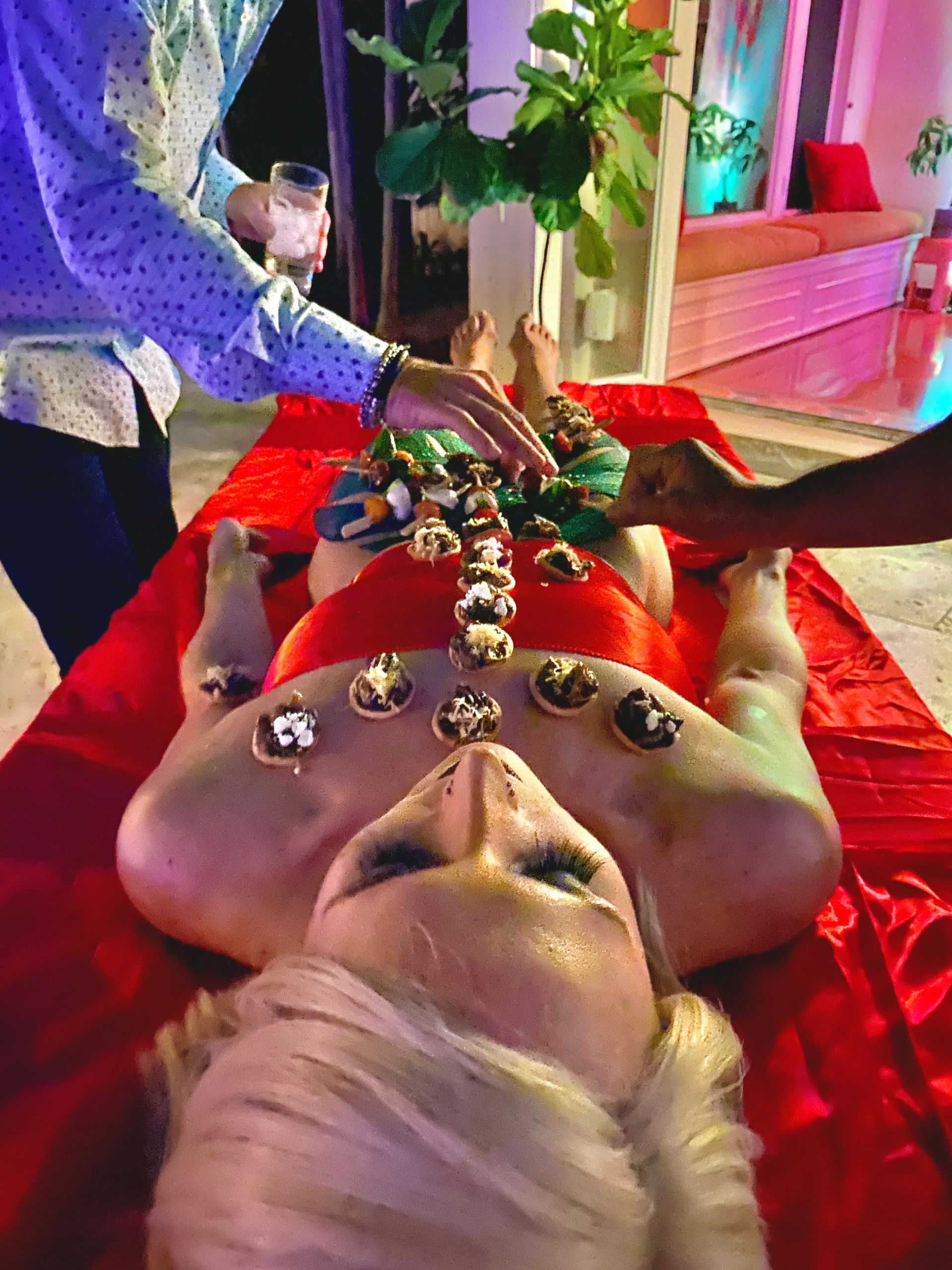 Person reclining on red satin cloth at a party while guests place bite-sized desserts along their torso, colorful mood lighting and indoor plants in the background