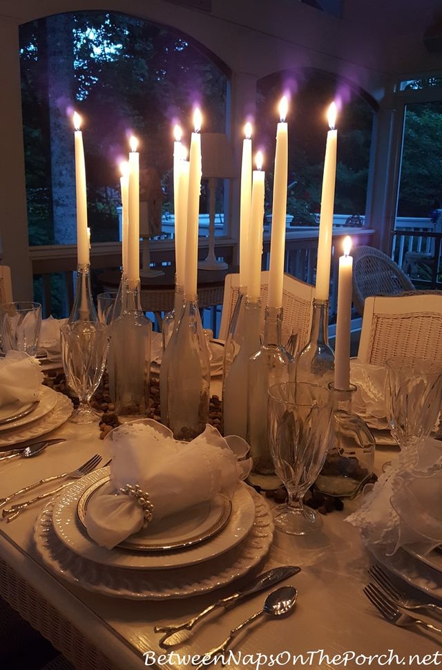 Candlelit dinner table on a screened porch at dusk — tall taper candles in clear glass bottles, white china, crystal goblets, pearl napkin rings, and silver flatware.