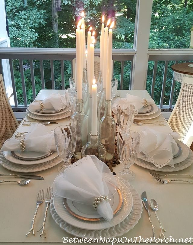 Elegant white tablescape on a screened porch set for six with layered white plates, pearl napkin rings, crystal goblets, and tall taper candles in glass bottles overlooking a leafy green backyard