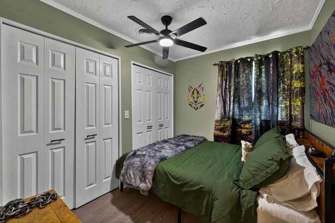 Cozy bedroom with olive-green walls and green bedding, faux-fur gray throw, forest-print curtains, stylized wolf wall decal, black ceiling fan, white double closet doors and hardwood floor.