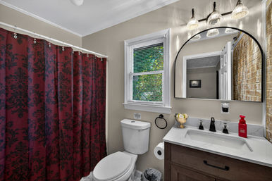 Bright residential bathroom with maroon damask shower curtain, white toilet, marble-look vanity, arched mirror, black fixtures and brick accent wall