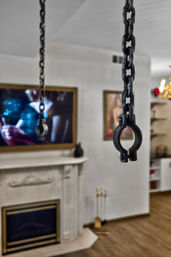 Close-up of black metal chains and a round clamp hanging in a modern living room interior with a marble fireplace, wall-mounted TV and framed artwork blurred in the background.