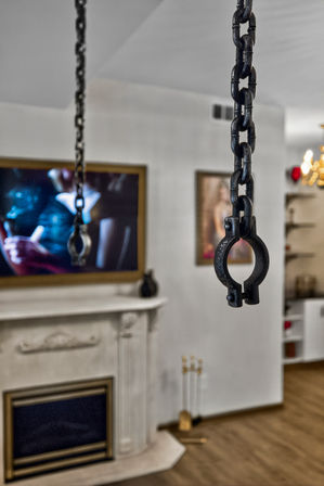 Close-up of black metal chains and a round clamp hanging in a modern living room interior with a marble fireplace, wall-mounted TV and framed artwork blurred in the background.