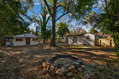 Shaded suburban backyard with a large oak tree, circular stone fire pit in the foreground, a small white outbuilding with a rooftop sign at left, and a single-story house with an arched window and wooden fence at right, surrounded by leafy trees and dappled sunlight.