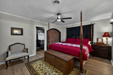 Cozy vintage-style bedroom with a dark wood four-poster bed dressed in deep red bedding and floral runner, carved wooden chest at the foot, patterned area rug, beige upholstered armchair, dark curtains, wooden nightstand with red lamp, and modern ceiling fan.