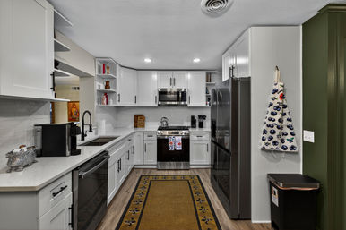 Bright modern white kitchen with stainless-steel appliances, black fixtures, farmhouse sink, wood-look floors, patterned runner rug and hanging blueberry apron.