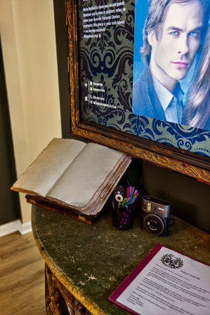 Vintage guestbook open on a wooden stand beside a framed digital display with a portrait, pen cup of colorful markers and an instant camera on a rustic hotel lobby reception table.